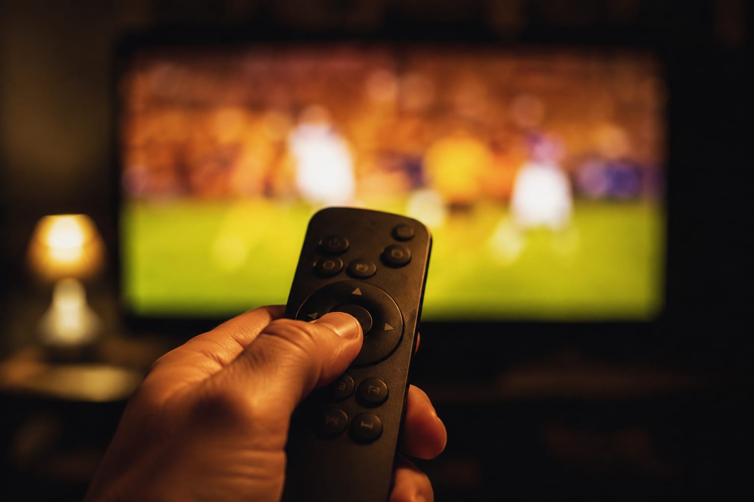 A hand holding a streaming remote in front of a television showing a channel guide