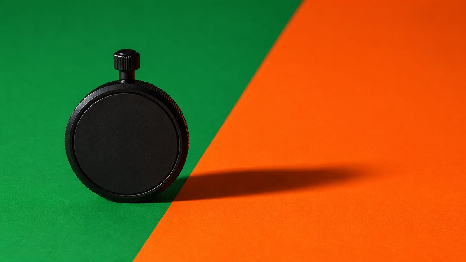 A matte-black stopwatch standing upright on a backdrop split diagonally from stadium green into hot orange