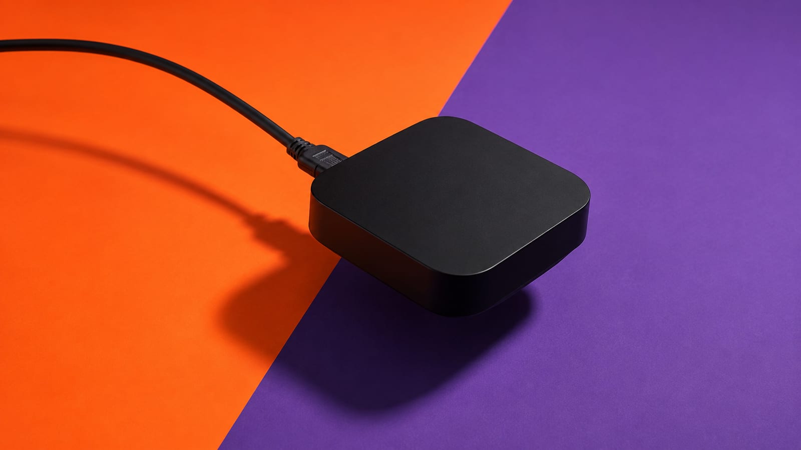 A black cable plugged into a matte-black streaming box on a hard diagonal split of orange and violet