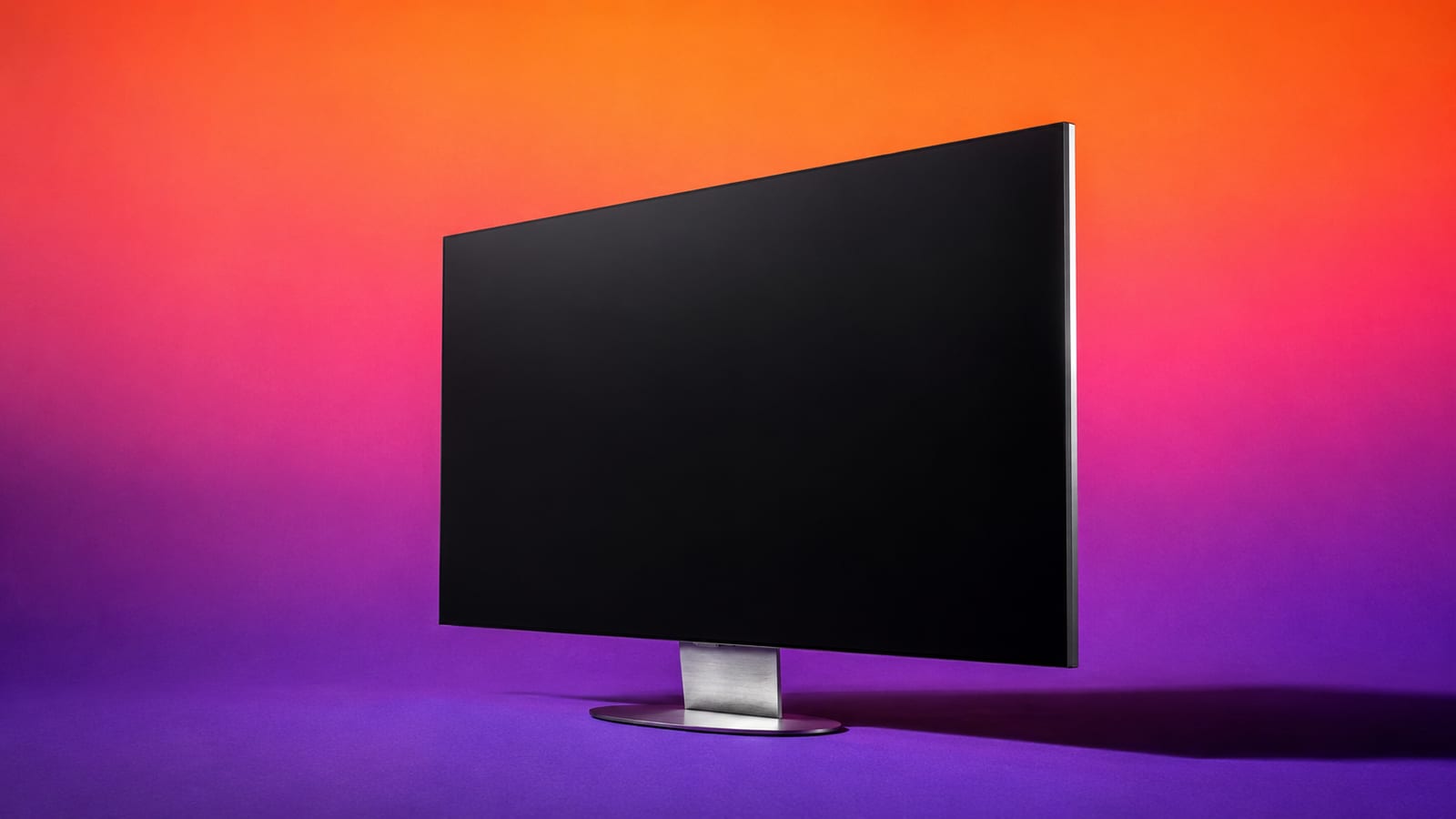 A black flat-screen television on a slim brushed-metal stand against a saturated orange and violet backdrop