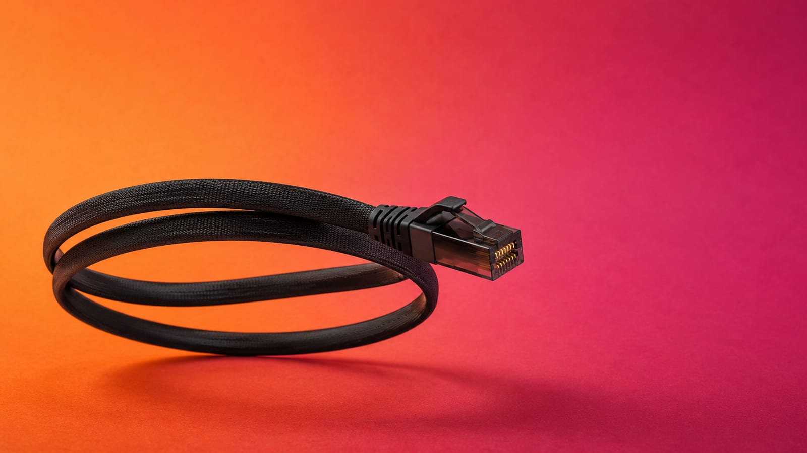 A flat black ethernet cable coiled in mid-air with its RJ45 connector facing the camera, on an orange and magenta backdrop
