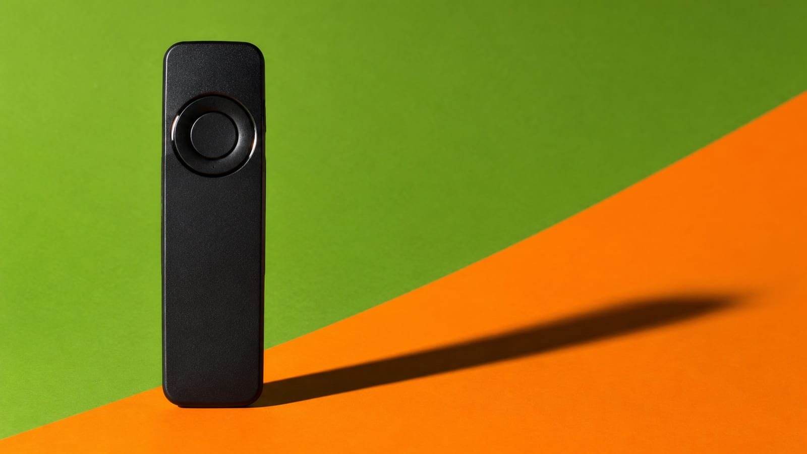 A slim black television remote standing upright on a backdrop split diagonally from stadium green into hot orange