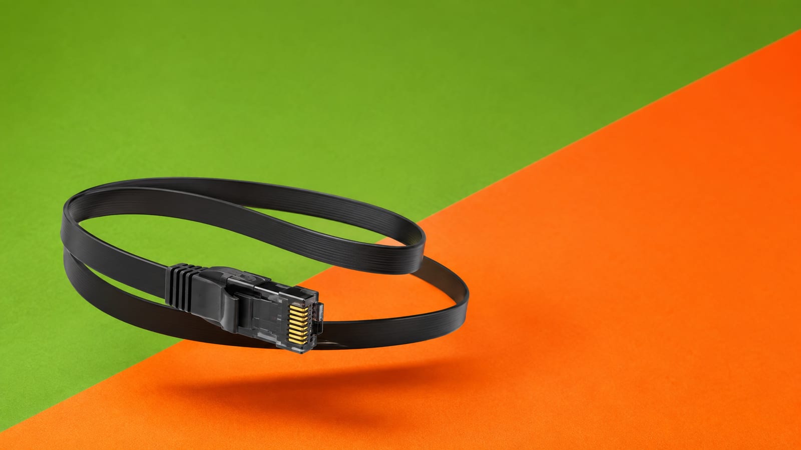 Flat matte-black ethernet cable coiled in the air with its connector turned to camera on a green and orange split backdrop