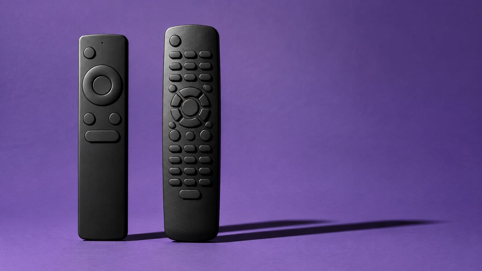 A simple television remote beside an older button-heavy remote on a violet studio background