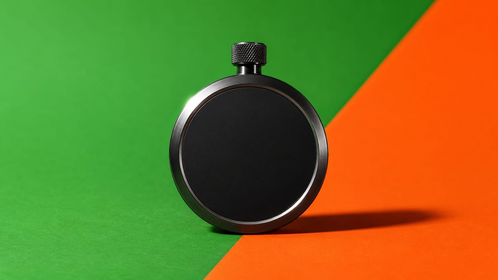 A matte-black stopwatch standing upright on a backdrop split diagonally from stadium green into hot orange